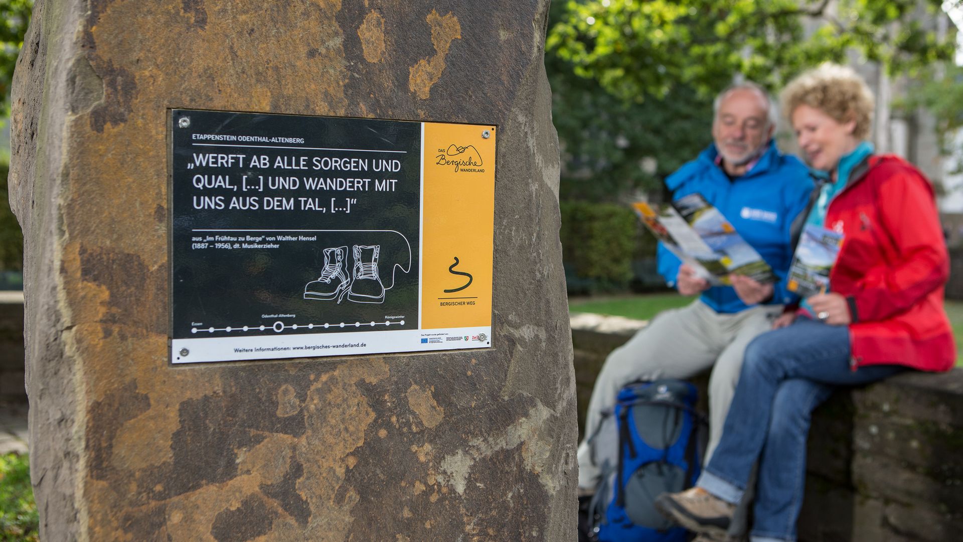 Vorne ist ein ca. 1,5 m hoher Grauwackestein mit einem Infoschild sichtbar. Rechts daneben ist im Hintergrund unscharf ein Paar in Wanderkleidung sichtbar. 