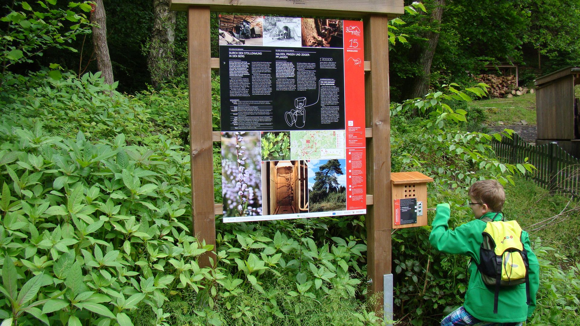 Inmitten grüner Sträucher steht eine Infotafel des Bergbauweges. An der daran angebrachten Audiostation kurbelt ein Kind in grüner Jacke und mit gelbem Rucksack an der Kurbel.